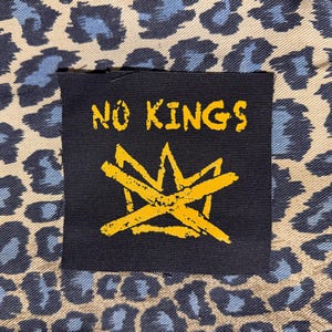 May include: A black square patch with the words "NO KINGS" in yellow, above a yellow crown design. The patch is set against a background of tan fabric with blue leopard print.