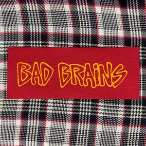 May include: A rectangular red fabric patch featuring the yellow text BAD BRAINS in a stylized font, stitched onto a black, white, and red plaid fabric background. The patch is centered horizontally and vertically against the patterned textile.