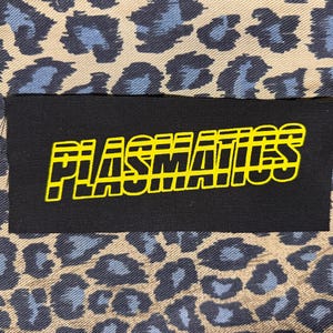 May include: A rectangular black fabric patch featuring the word PLASMATICS in a bold, yellow, horizontally-striped font. The patch is centered over a tan and blue leopard print fabric background with a repeating animal spot pattern.