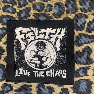 May include: A black patch with white text and a graphic of a person in a circle, set against a leopard print background. The patch reads "FILTH PUNKS" and "LIVE THE CHAOS".