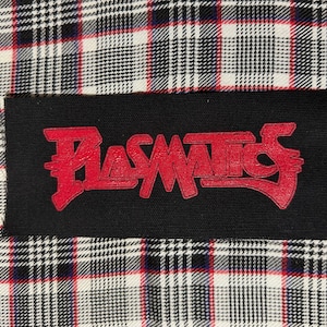 May include: A rectangular black fabric patch featuring the word Plasmatics embroidered in bold red lettering. The patch is positioned against a background of white, black, and red plaid fabric with a classic woven textile pattern throughout.