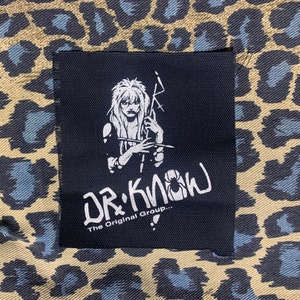 May include: Black and white patch with a drawing of a woman holding a microphone and a lightning bolt. The text "Dr. Know The Original Group..." is below the drawing.