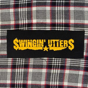 May include: A rectangular black fabric patch featuring the yellow text Swingin' Utters with dollar signs on each end and a small star in the center, sewn onto a white, black, and red plaid fabric background with a visible woven textile pattern.