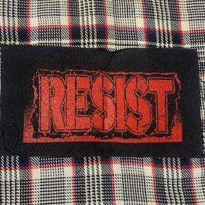 May include: A black fabric patch with the word "RESIST" printed in red letters. The patch has a distressed, vintage look.