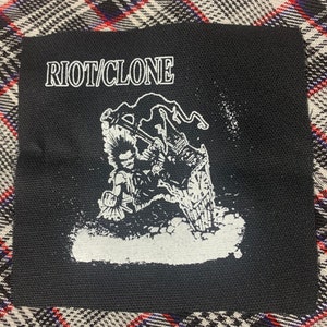 May include: Black square patch with the text "RIOT/CLONE" in white, above a white graphic of a punk rocker and a clock tower. The patch has a distressed, worn look.