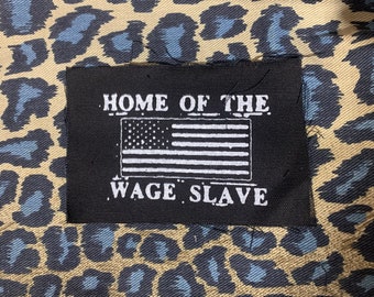 May include: Black rectangular patch with white text and an American flag graphic. The text reads "HOME OF THE WAGE SLAVE." The patch is set against a tan and blue leopard print background.
