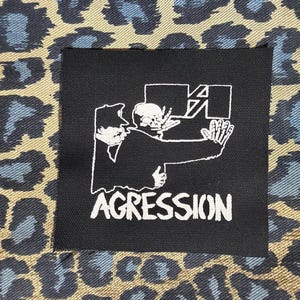 May include: A black square patch with the word "AGGRESSION" in white, featuring a white graphic of two figures, one a skeleton, in a confrontational pose. The background is a tan and blue leopard print.
