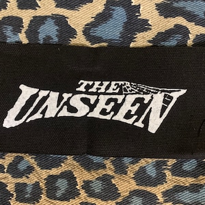 May include: Black and white fabric patch with the text "THE UNSEEN" in a bold, stylized font.