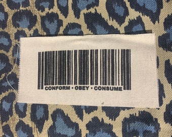 May include: A white fabric patch with a black barcode and the text "CONFORM OBEY CONSUME" printed on it.