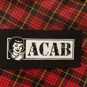 May include: Black and white patch with a woman's face and the text "ACAB" in a white rectangle.