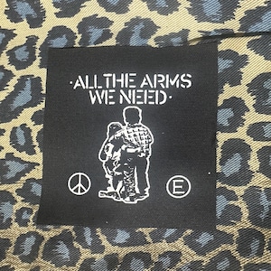 May include: Black and white patch with the text "ALL THE ARMS WE NEED." and a graphic of two people hugging. The patch has a peace sign and a letter "E" on either side of the graphic.