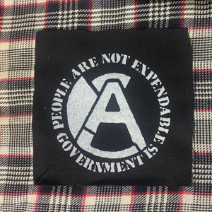 May include: Black patch with white text that reads "People are not expendable, government is". The text is in a circle with an "A" in the center.
