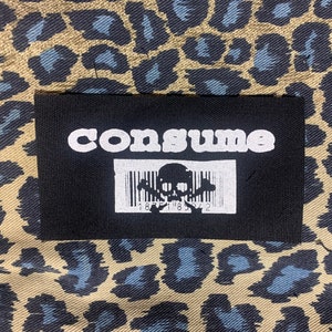 May include: Black patch with white text that reads "consume" and a skull and crossbones graphic with a barcode underneath. The patch is on a brown and blue leopard print fabric.