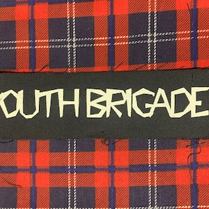 May include: Black patch with white text that reads "YOUTH BRIGADE" on a red and blue plaid background.