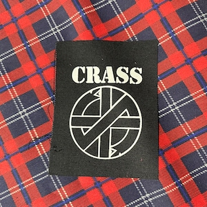 May include: Black and white patch with the word "CRASS" in white letters and a circle with a diagonal line through it. The patch is on a red and blue plaid background.