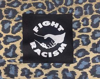 May include: Black patch with white text that reads "Fight Racism" and a white silhouette of two hands shaking. The patch is on a brown and blue leopard print fabric.