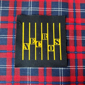May include: Black patch with yellow vertical lines and the word "NECROS" written vertically in yellow letters.
