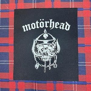 May include: A square black fabric patch featuring the white printed text motörhead above a detailed white graphic of a snarling warthog-like creature wearing a spiked helmet, chains, and a cross pendant, centered on a red and blue plaid background.