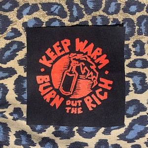 May include: A black patch with red text that reads "Keep Warm. Burn Out The Rich."  The text is in a circle with a red hand holding a spray bottle inside the circle.