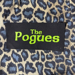 May include: A black patch with the text "The Pogues" in green lettering on a leopard print fabric background.