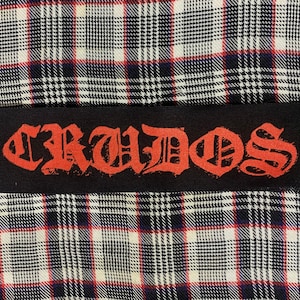May include: A black patch with the word "CRADOS" printed in red, gothic-style lettering. The patch is sewn onto a plaid fabric background.