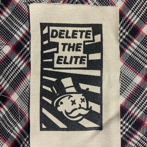 May include: A black and white patch with the text "DELETE THE ELITE" and a cartoon image of a person with a top hat and an X over their eyes.