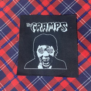 May include: Black and white iron-on patch with the text "The Cramps" and a graphic of a person with a skull-like face wearing goggles.