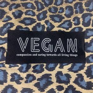 May include: A black patch with white text that reads "VEGAN" and the phrase "compassion and caring towards all living things". The patch is on a fabric background with a leopard print pattern.
