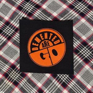 May include: A square black fabric patch featuring a circular orange graphic with the text DEMENTED ARE GO printed in bold letters. The patch is centered on a white, black, and red plaid fabric background with a visible woven texture throughout.