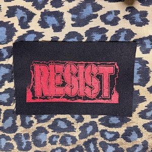 May include: A black rectangular patch with the word "RESIST" in red, distressed lettering. The patch is set against a background of tan fabric with blue and black leopard print.