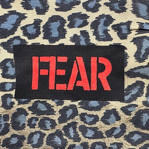 May include: A black patch with the word "FEAR" in red letters on a beige background with a blue leopard print pattern.