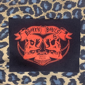 May include: Red and black embroidered patch with two skulls and the text "BROKEN BONES!"