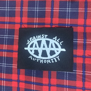 May include: Black patch with white text and graphic. The text reads "Against All Authority" and the graphic is a circle with the letters "AAA" inside.
