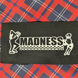 May include: Black rectangular patch with the word "MADNESS" in white block letters. The patch features two white cartoon figures, one on each side of the word, and a checkered pattern beneath the word.