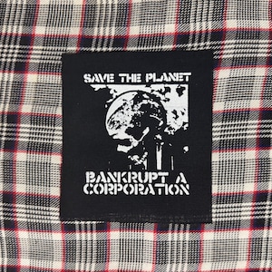 May include: A black rectangular fabric patch featuring a white graphic of a globe and the text "SAVE THE PLANET" and "BANKRUPT A CORPORATION" stitched onto a white, black, and red plaid fabric background with a classic woven tartan pattern.