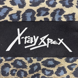 May include: A black rectangular label with the white text "X-ray Spex" is set against a leopard print background in tan and blue. The leopard print has a repeating pattern of blue spots.