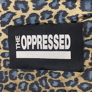 May include: A black patch with white text that reads "THE OPPRESSED" on a beige and blue leopard print fabric.