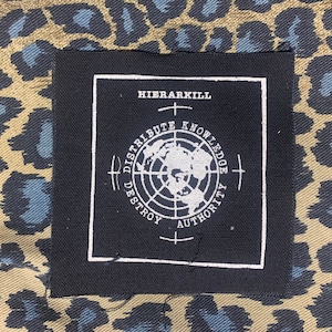 May include: A black square patch with white text and a world map graphic. The text reads "HIERARKILL", "DISTRIBUTE KNOWLEDGE", and "DESTROY AUTHORITY". The patch is set against a tan and blue leopard print background.