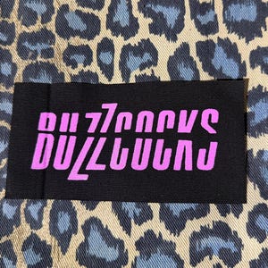 May include: A black rectangular patch with the word "BUZZCOCKS" in bright pink, stylized lettering. The patch is set against a background of tan and blue leopard print fabric.