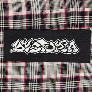 May include: A black patch with the white word "Dystopia" in a graffiti-style font. The patch is rectangular and set against a plaid fabric background with red, white, and black colors.