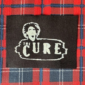 May include: A black rectangular patch featuring the band name "THE CURE" in white, with a stylized image of a person's face above the text. The patch is set against a red, blue, and white plaid background.
