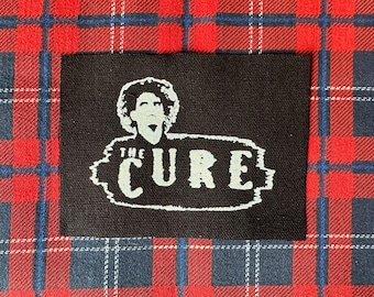May include: A black rectangular patch featuring the band name "THE CURE" in white, with a stylized image of a person's face above the text. The patch is set against a red, blue, and white plaid background.