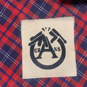 May include: A black and white patch with the word "GRASS" and a stylized letter "A" in a circle. The letter "A" is formed by two hands holding a gun.