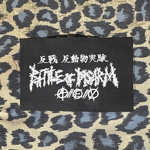 May include: A black patch with white text that reads "反戦 反動 物実質" and "BATTLE OF PSYCHO" with a circle containing an anarchy symbol on either side. The patch is on a brown and blue leopard print fabric.