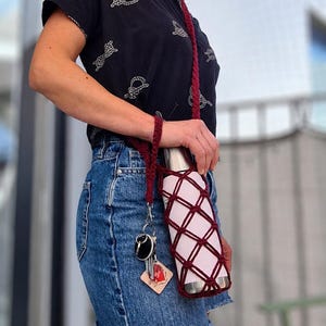 May include: A maroon crocheted water bottle holder with a diamond-patterned net design and a long braided strap. The holder features a small metal clip attached to a set of keys and a square wooden tag with a red heart graphic and text.