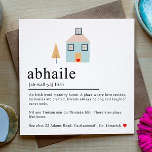 May include: A square greeting card featuring the Irish word abhaile, a graphic of a house with a small red heart, and text defining the word as home. The card is placed on a brown envelope, surrounded by bright pink flowers and two heart-shaped objects.