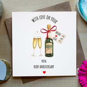 Op de afbeelding: Een wenskaart met een illustratie van een champagnefles en twee glazen, met de tekst "WITH LOVE ON YOUR 40TH RUBY ANNIVERSARY". De kaart is omgeven door bloemen en hartvormige decoraties.