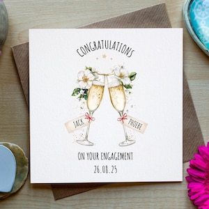 Op de afbeelding: Een vierkante verlovingskaart met de woorden "Gefeliciteerd" en "Op je verloving" erop gedrukt. De kaart toont illustraties van champagneglazen, bloemen en de namen "Jack" en "Phoebe". Omringd door bloemen en decoratieve harten.