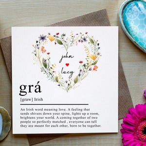 Könnte beinhalten: Eine Grußkarte mit dem irischen Wort "grá" für Liebe, umgeben von einem floralen Herzdesign. Die Karte enthält die Namen "John" und "Lucy". Das Bild zeigt auch Blumen und dekorative Herzen.