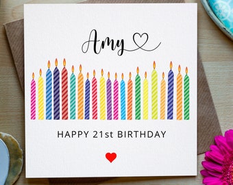 May include: A white card with the name "Amy" written in cursive with a small heart symbol. The card has a row of 21 colorful candles with flames on top. The text "HAPPY 21st BIRTHDAY" is printed below the candles. A small red heart is printed below the text.